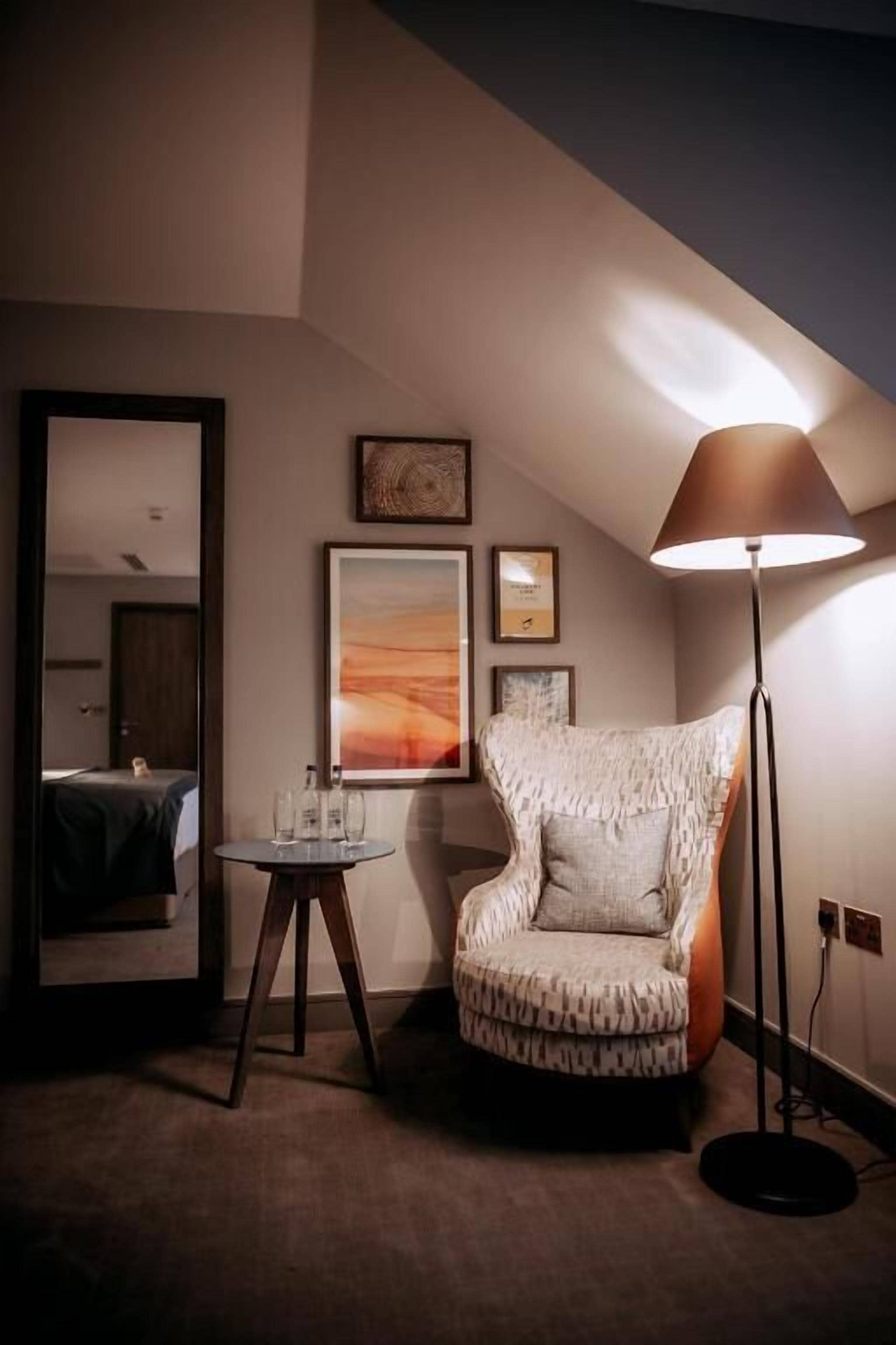 A hotel room with a patterned armchair, a standing lamp, a small round table, and framed artwork on the wall.