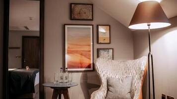 A hotel room with a patterned armchair, a standing lamp, a small round table, and framed artwork on the wall.