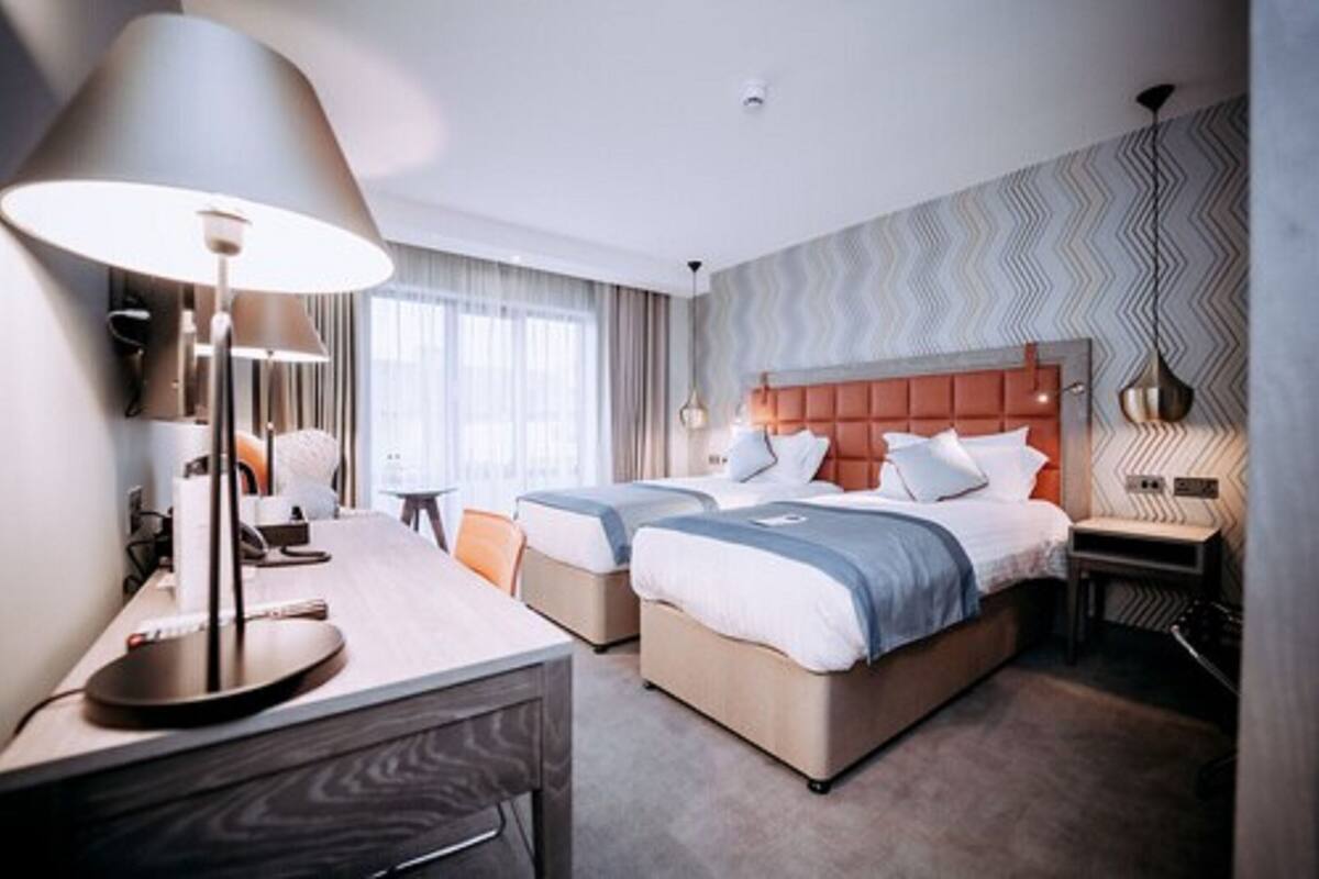 Ein Hotelzimmer mit zwei Betten, einem Schreibtisch, einer Lampe und Blick auf die Stadt.