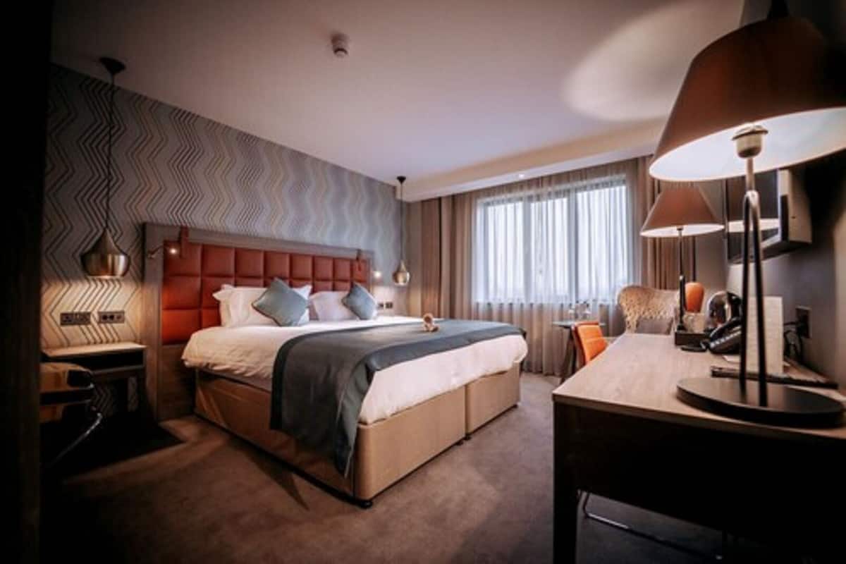 Ein Hotelzimmer mit einem großen Bett, einem Schreibtisch, einem Stuhl, einer Lampe und einem Fenster mit Vorhängen.