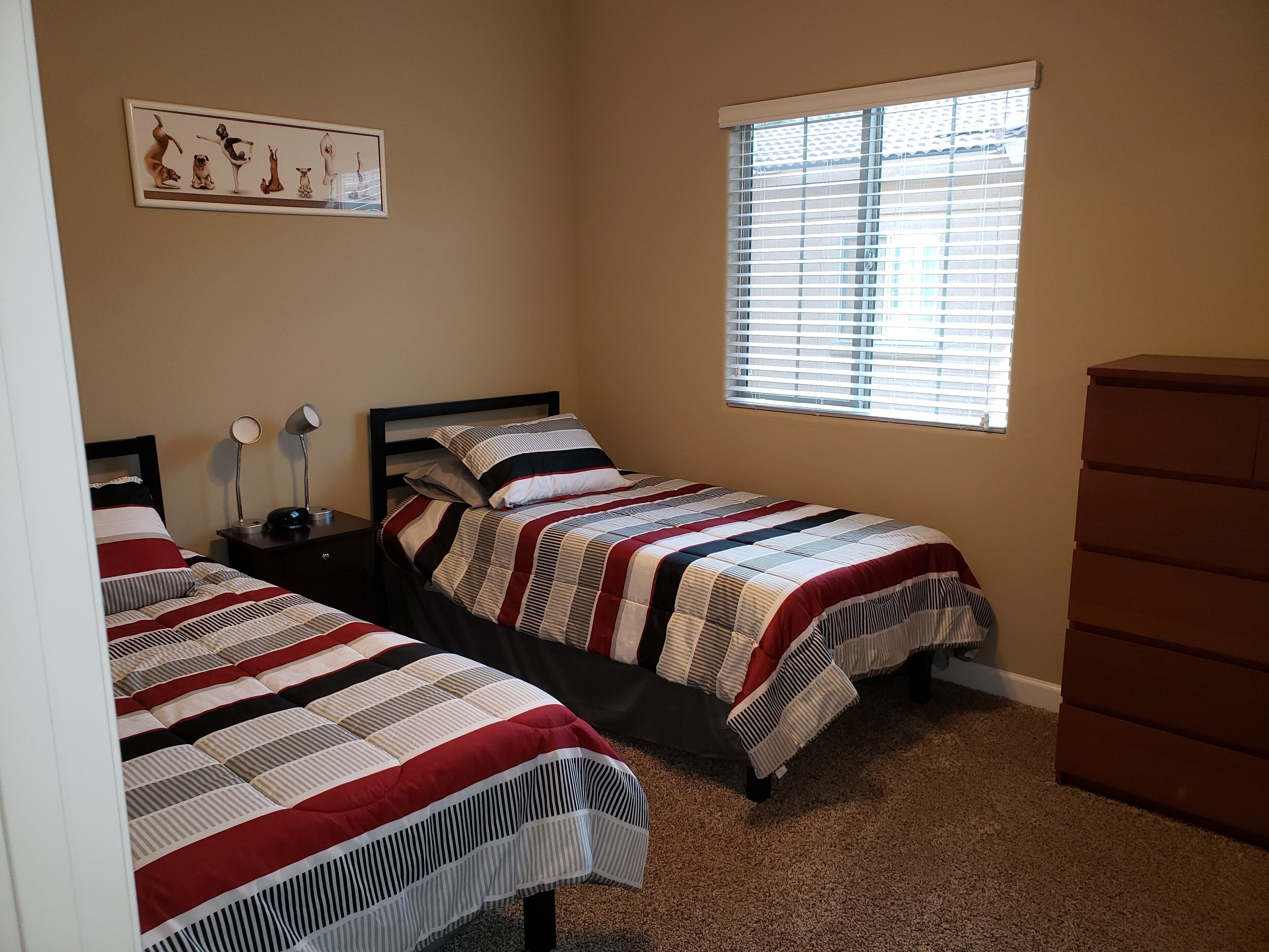 A bedroom with two beds, a dresser, a window with blinds, and a wall-mounted picture.