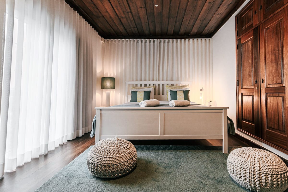 A bedroom with a wooden ceiling, a bed with two pillows, a bedside lamp, and a large wooden wardrobe.