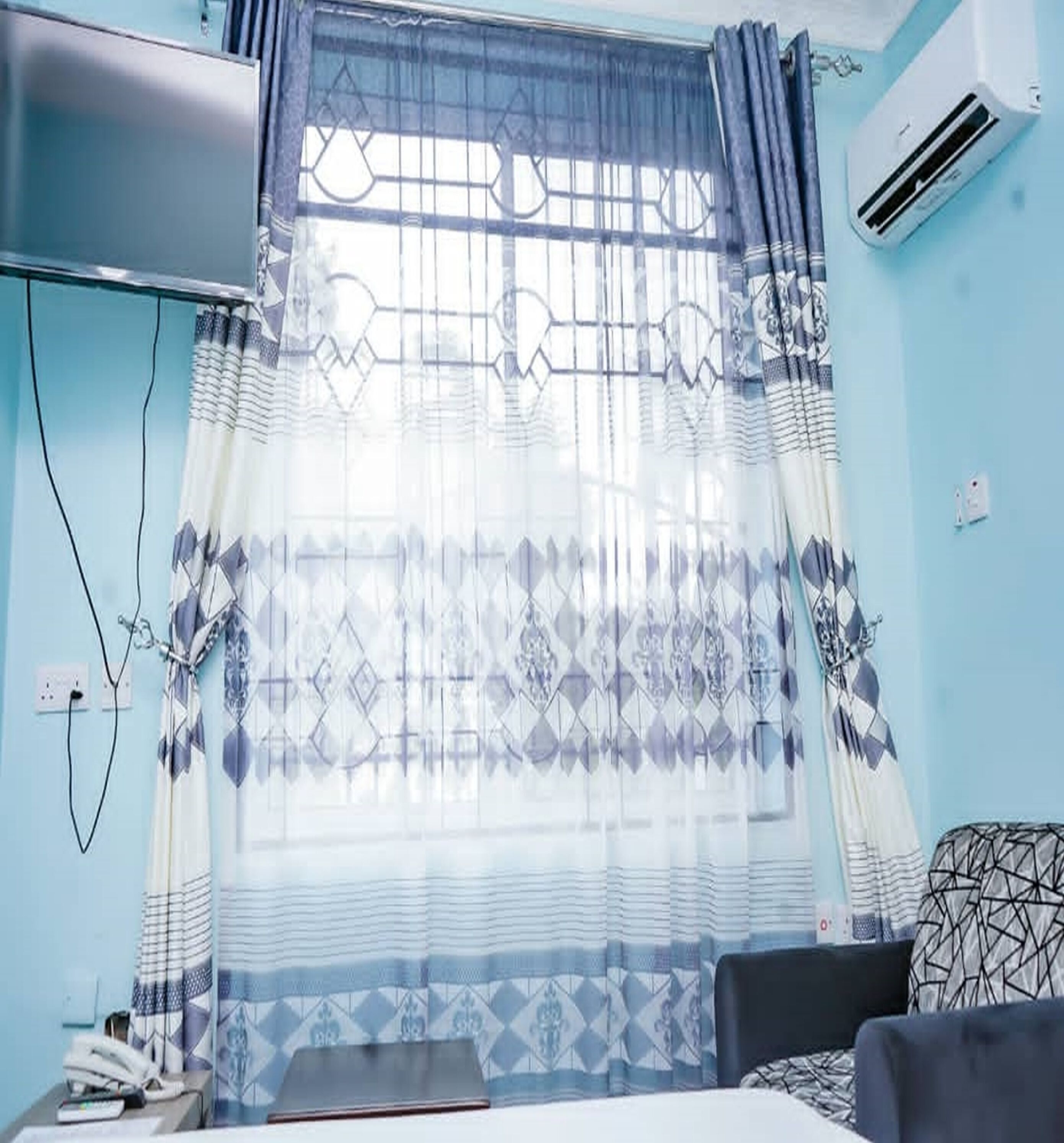 A hotel room with a bed, a bedside table with a phone and a book, a window with patterned curtains, and an air conditioning unit on the wall.