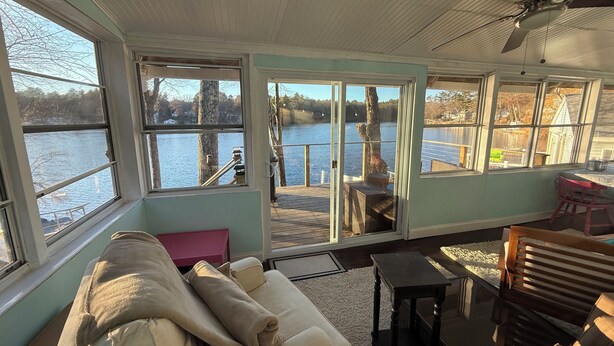 A cozy room with a large window overlooking a lake, a ceiling fan, and a wooden deck outside. - Your Cottage on the Lake (Dudley)