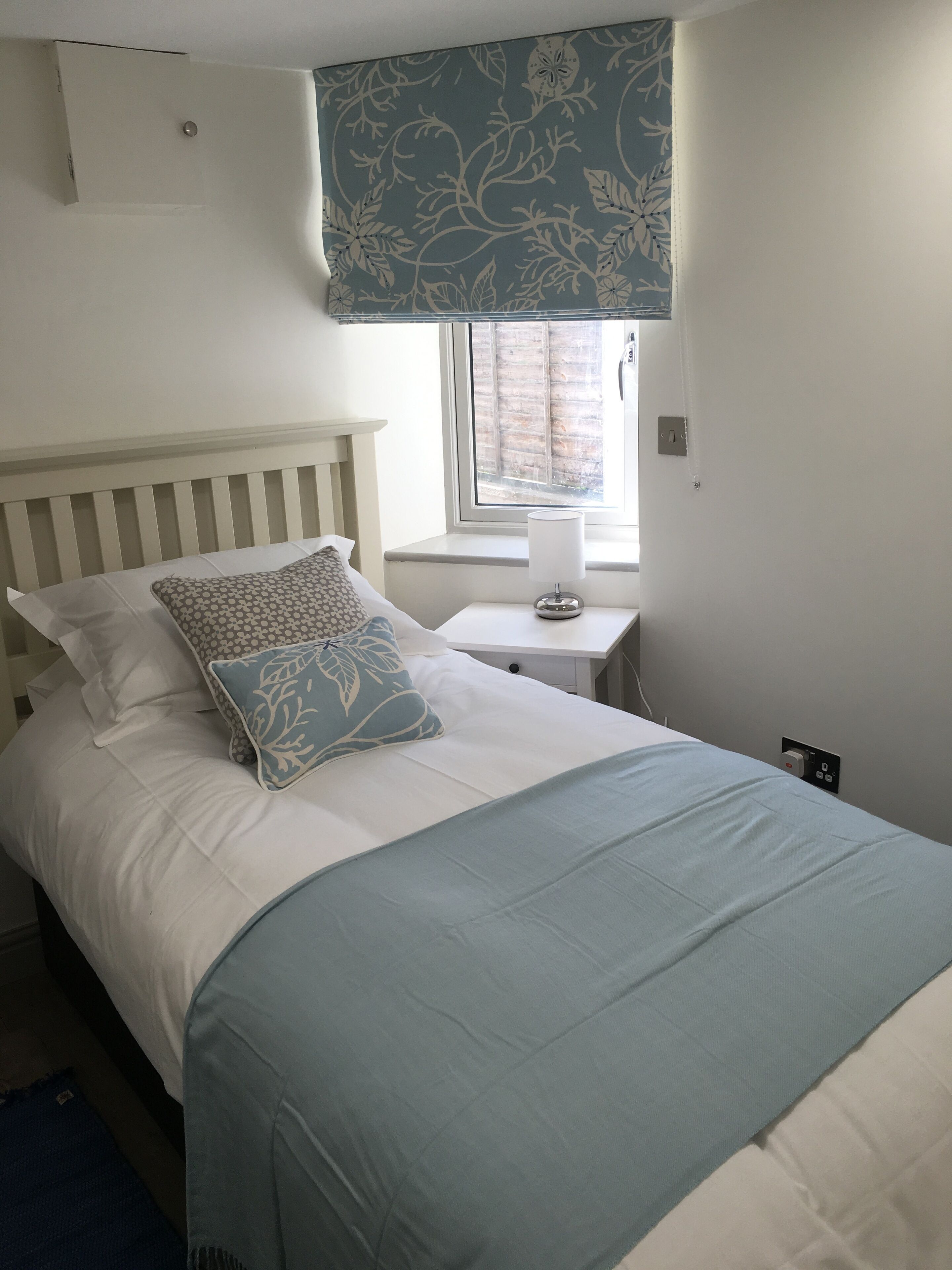 A neatly made bed with a white headboard, a blue and white patterned pillow, and a light blue bedspread. There is a small bedside table with a lamp and a power outlet on the wall.