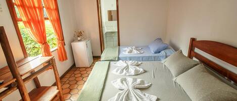 A room with two beds, a wooden desk, a small white cabinet, and a window with orange curtains.