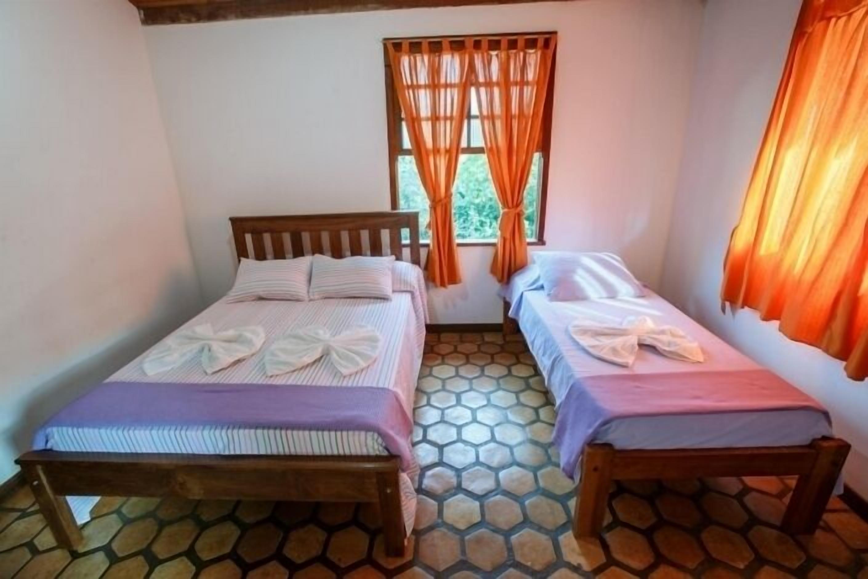 A room with two beds, each with a wooden headboard, and a window with orange curtains.