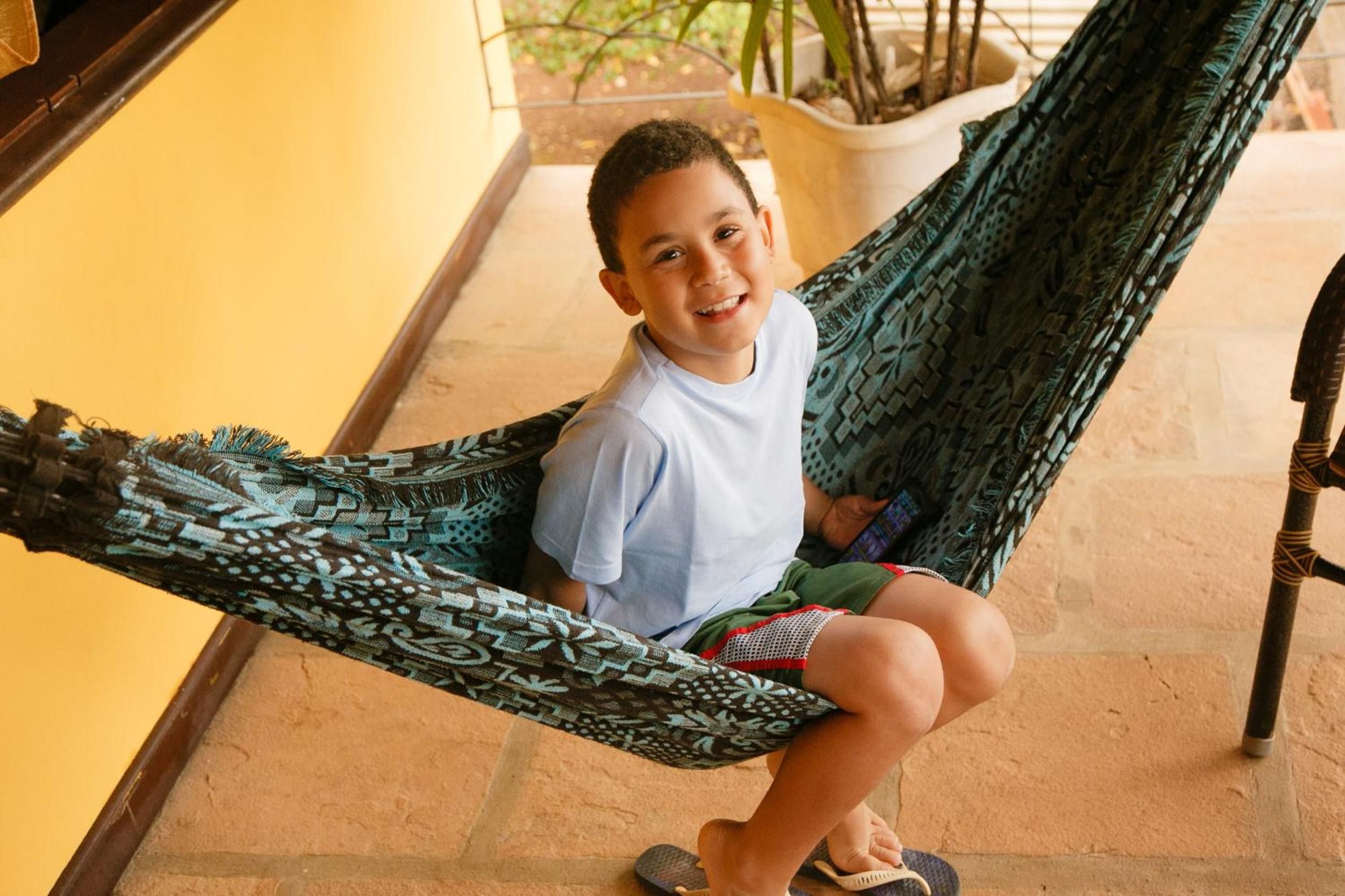 A person sitting in a hammock on a tiled floor with a yellow wall and a potted plant in the background.