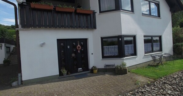 Ein zweistöckiges Haus mit einer schwarzen Tür, einer kleinen Veranda mit einer Bank und einem Gartenbereich mit Tisch und Stühlen.