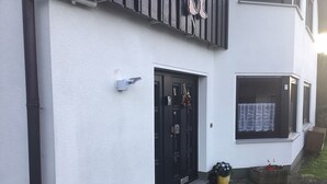 Un bâtiment blanc avec une porte noire, une petite fenêtre ornée d’un rideau et une boîte aux lettres noire fixée au mur.
