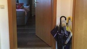 Un couloir mène à une chambre où se trouvent un lit, une table de chevet et une lampe. Une porte se trouve sur le côté droit, et un porte-parapluies contient plusieurs parapluies.