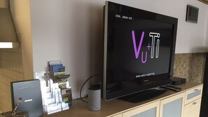 Une cuisine moderne équipée d’un grand téléviseur à écran plat affichant le logo « V+Ti », d’une machine à café, d’une bouilloire et d’un petit réfrigérateur.