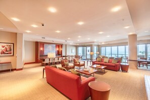 Un hall d’entrée spacieux, avec un canapé rouge, plusieurs tables et chaises, un comptoir d’accueil et de grandes fenêtres offrant une vue dégagée.