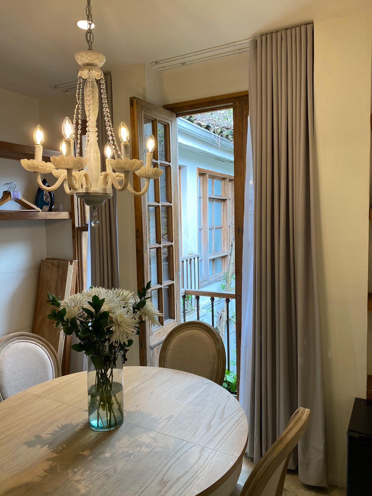 Un coin repas avec une table ronde, des chaises, un lustre, un vase de fleurs et une vue sur l’extérieur à travers une fenêtre ornée de rideaux.