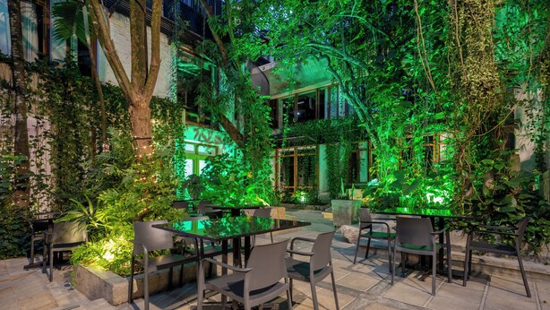 An outdoor patio with a glass table and chairs, surrounded by lush greenery and illuminated by green lighting. - Alko Hotel Casa Nispero (Cali)