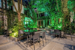 Un patio al aire libre con una mesa y sillas de vidrio, rodeado de exuberante follaje e iluminado con luces verdes.