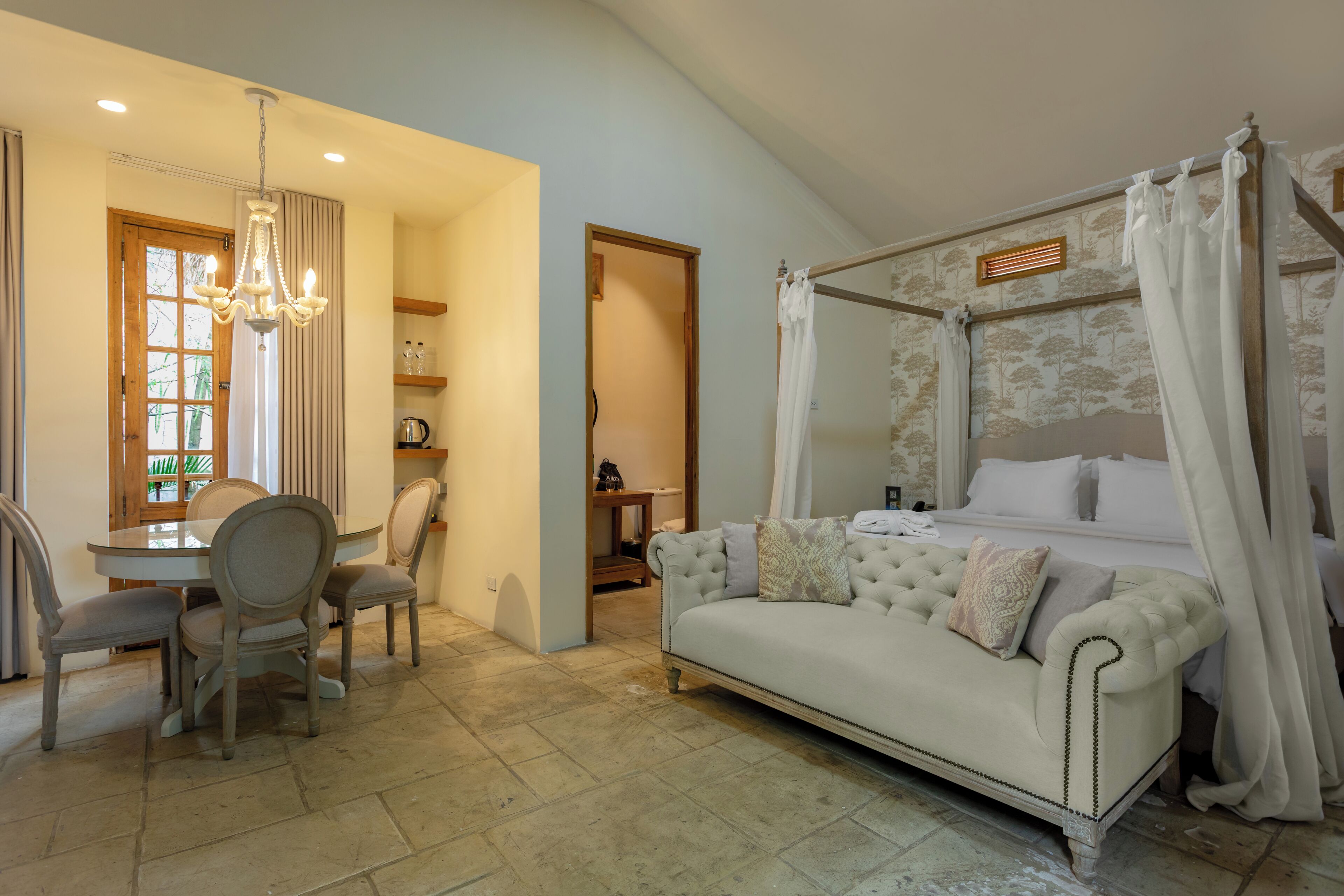 A bedroom with a four-poster bed, a sitting area with a sofa and chairs, a dining table with chairs, and a chandelier.