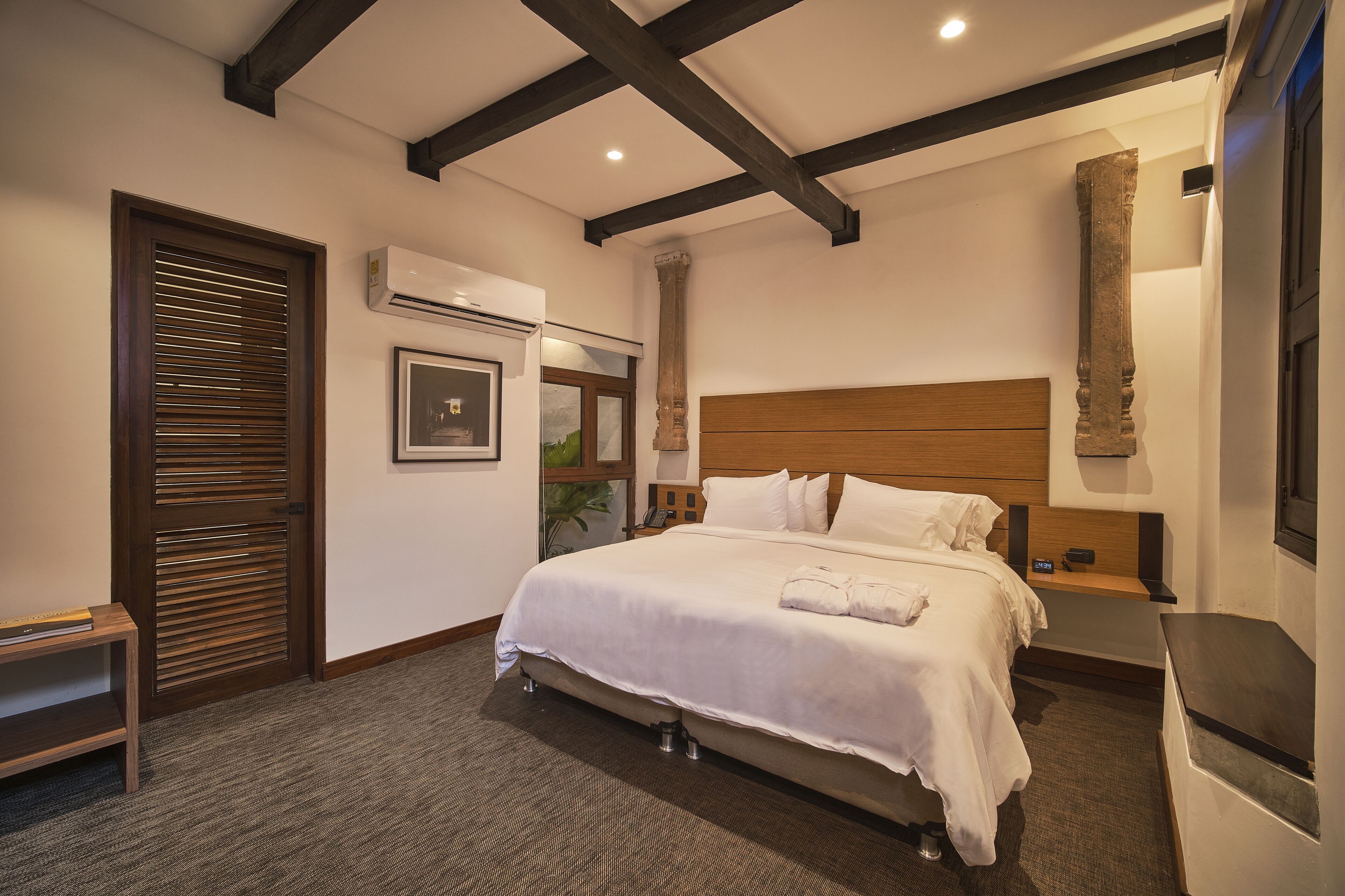 Una habitación de hotel con una cama grande, cabecero de madera, mesita de noche y aire acondicionado instalado en la pared.
