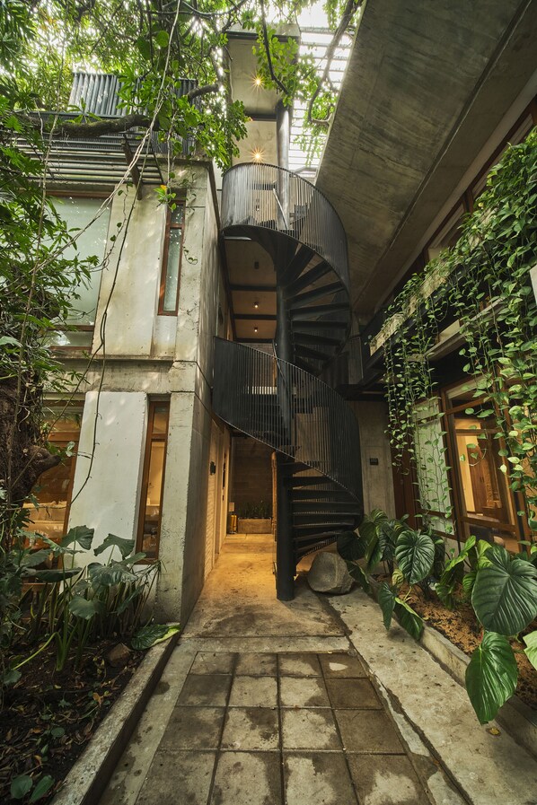 A spiral staircase in a courtyard surrounded by lush greenery and concrete structures.