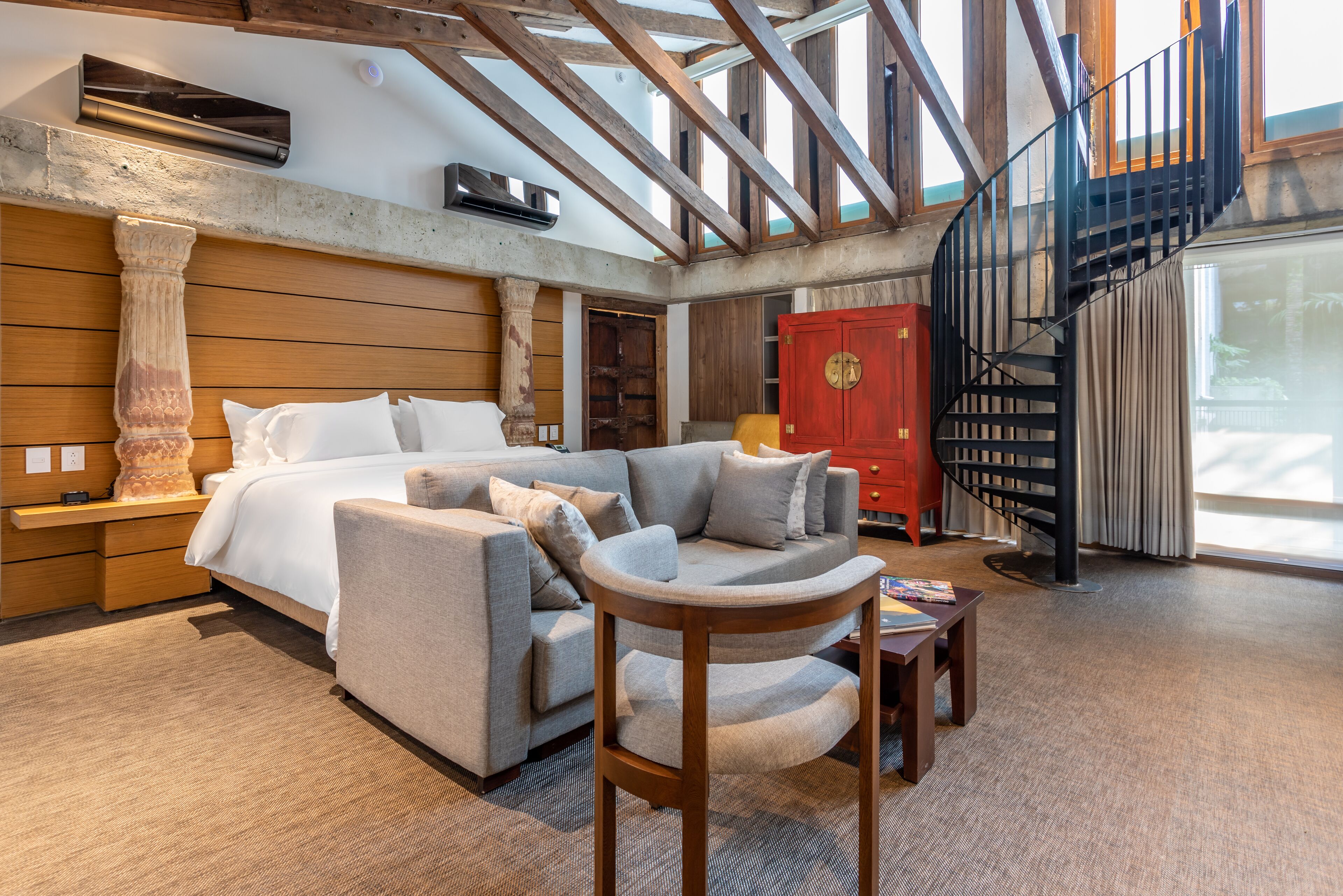A spacious living area with a large bed, a sofa, a wooden chair, and a red cabinet. There is a staircase with a black railing leading to an upper level.