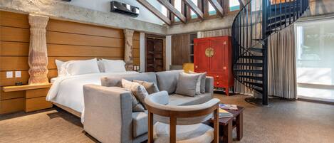 A spacious living area with a large bed, a sofa, a wooden chair, and a red cabinet. There is a staircase with a black railing leading to an upper level.