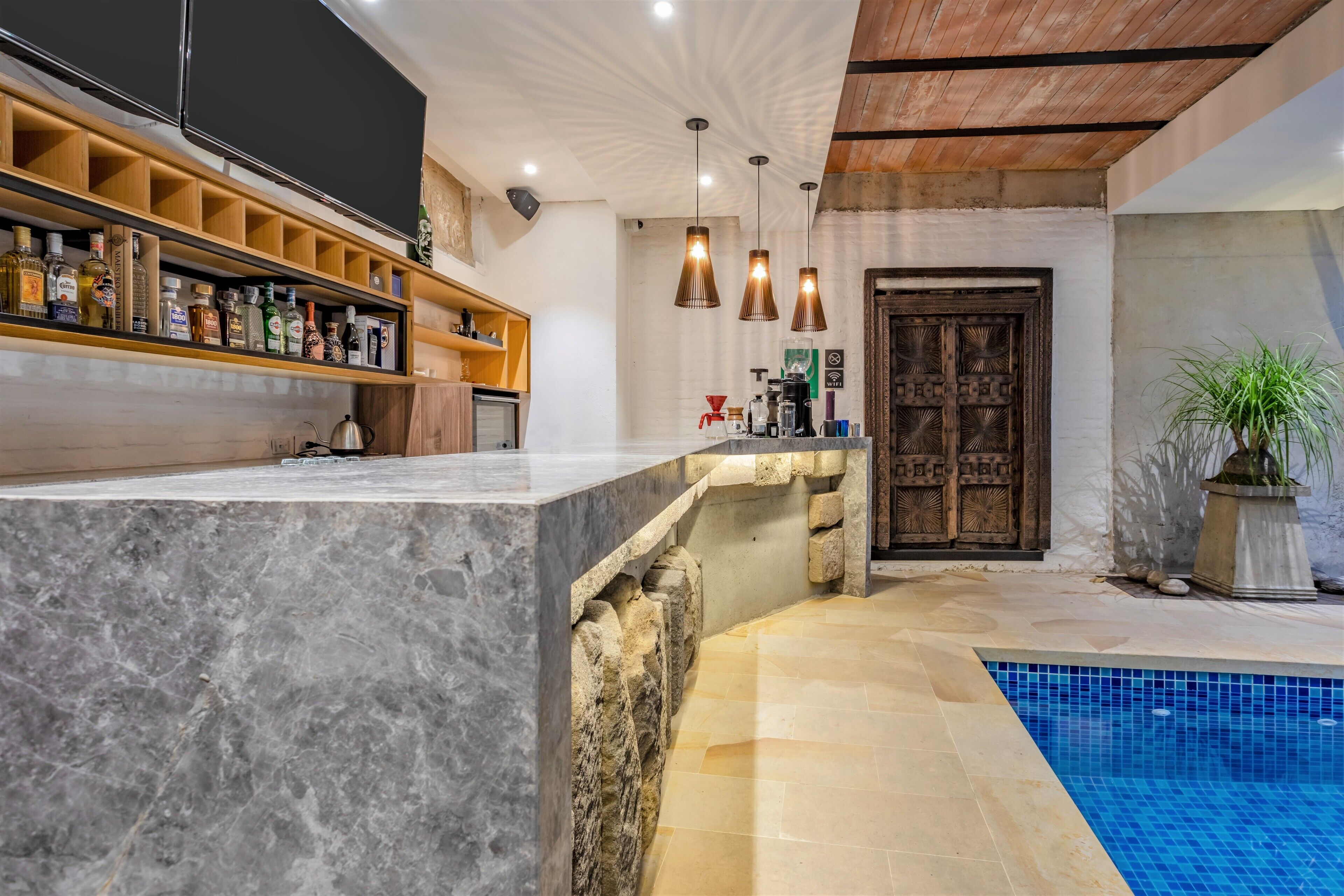 A modern kitchen with marble countertops, wooden shelves, hanging pendant lights, and a stone wall.