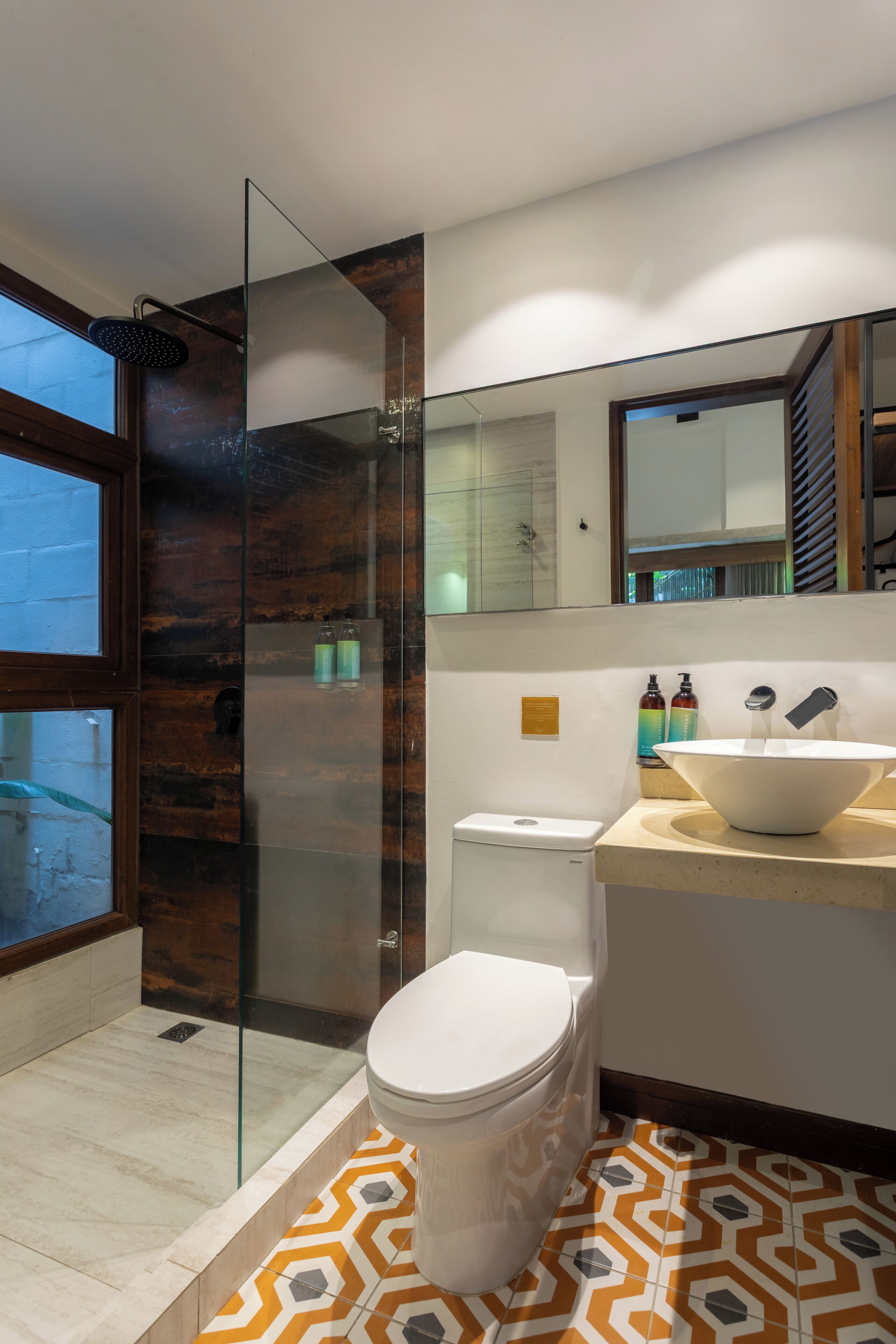 Une salle de bain moderne avec une douche à l’italienne en verre, des toilettes blanches, un grand lavabo sur colonne et un miroir au-dessus du lavabo.