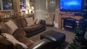 A living room with a sectional sofa, a black ottoman, a television, and a fireplace. - Macleods Guest House (Blackpool)