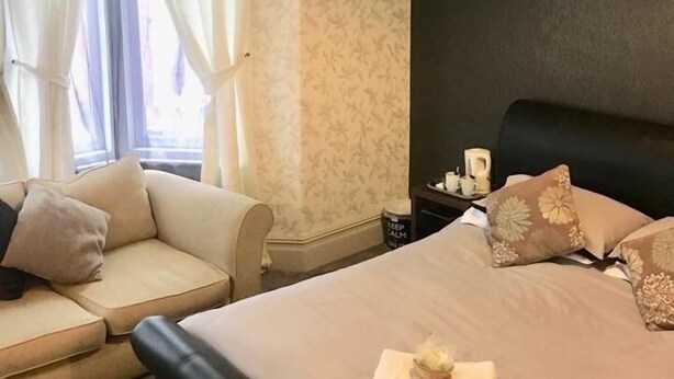 A hotel room with a bed, a sofa, a nightstand, and a window with curtains. - Macleods Guest House (Blackpool)