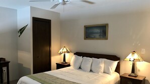 Una cama bien hecha con ropa de cama blanca y un plaid verde, flanqueada por dos mesitas de noche con lámparas, un cuadro en la pared y un ventilador de techo arriba.