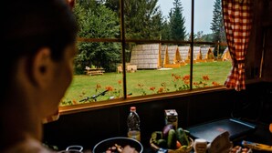 A room with a window overlooking a grassy area with wooden structures. There is a table with food items and a bottle. - Glamping FOREST EDGE (Kamnik)