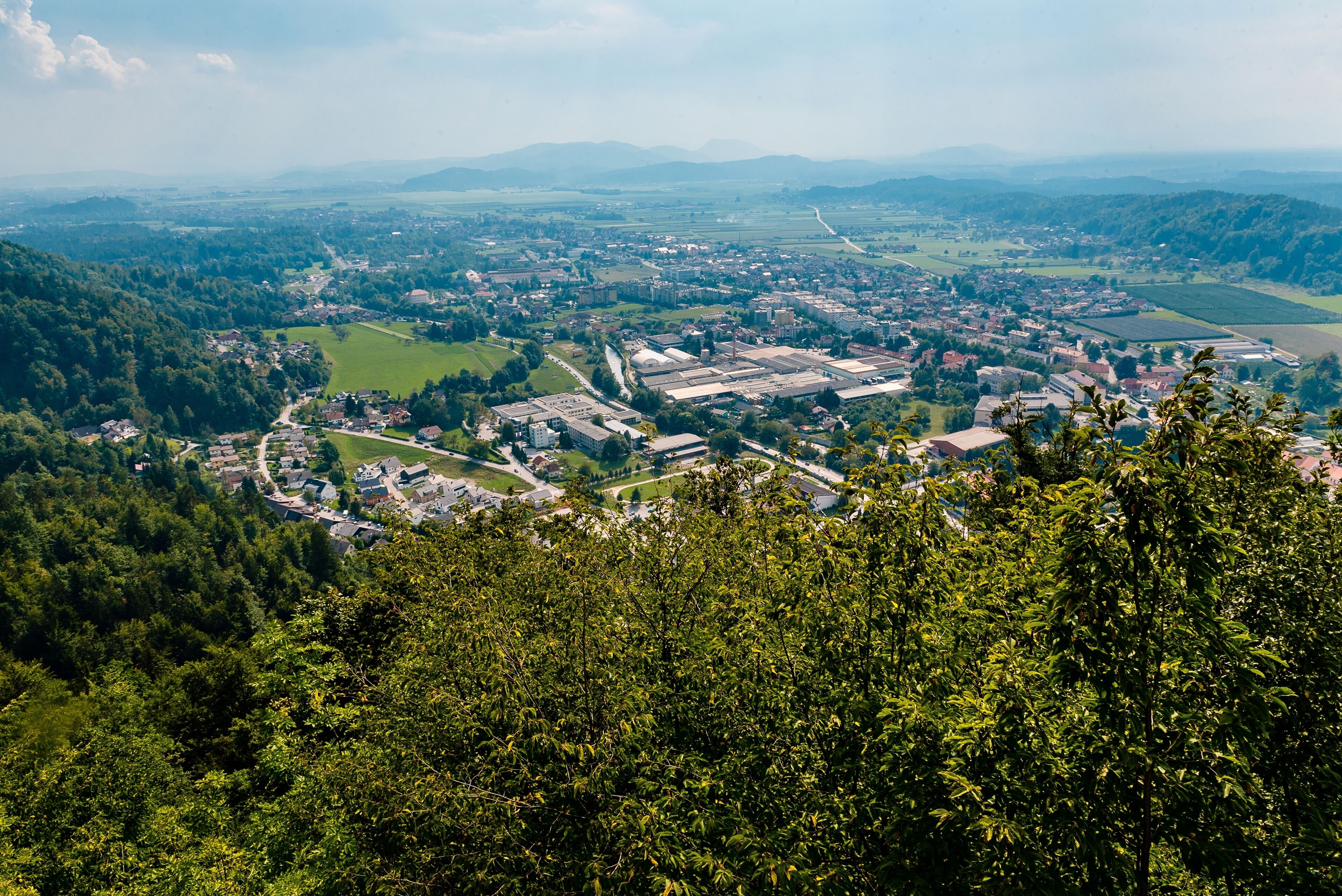 A town with a mix of residential and industrial buildings, surrounded by green hills and a clear sky.