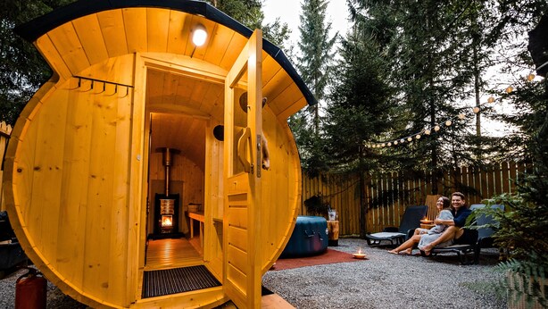 A wooden cabin with an open door, a bench, and a couple sitting outside. - Glamping FOREST EDGE (Kamnik)