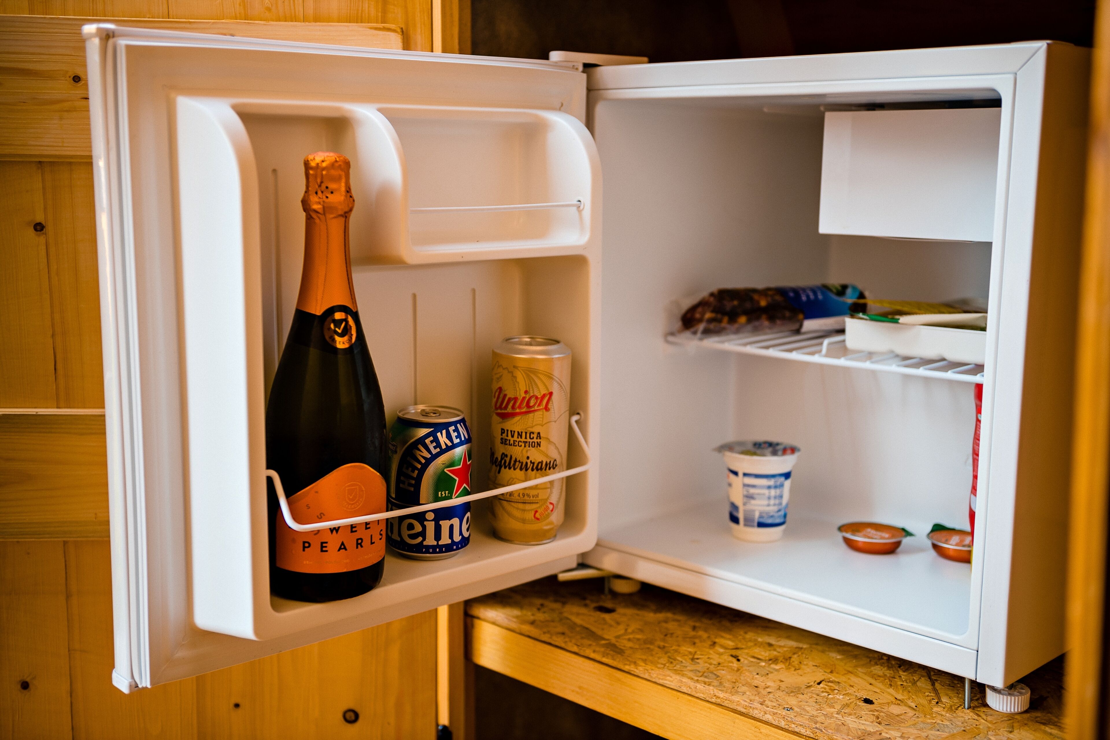 Un mini refrigerador con una botella de champán, una lata de cerveza y un recipiente con yogur.