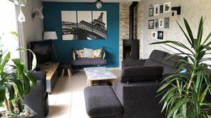 Een woonkamer met een bank, poef, salontafel en een open haard. Er zijn planten, een televisie en een schilderij aan de muur.
