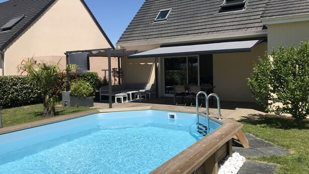 Zwembad met houten terras, een overdekte patio met buitenmeubilair en een huis met schuin dak.