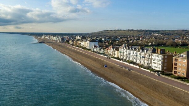 Un pueblo costero con una extensa playa arenosa, un paseo marítimo flanqueado por edificios y vistas al mar y colinas en la distancia.
