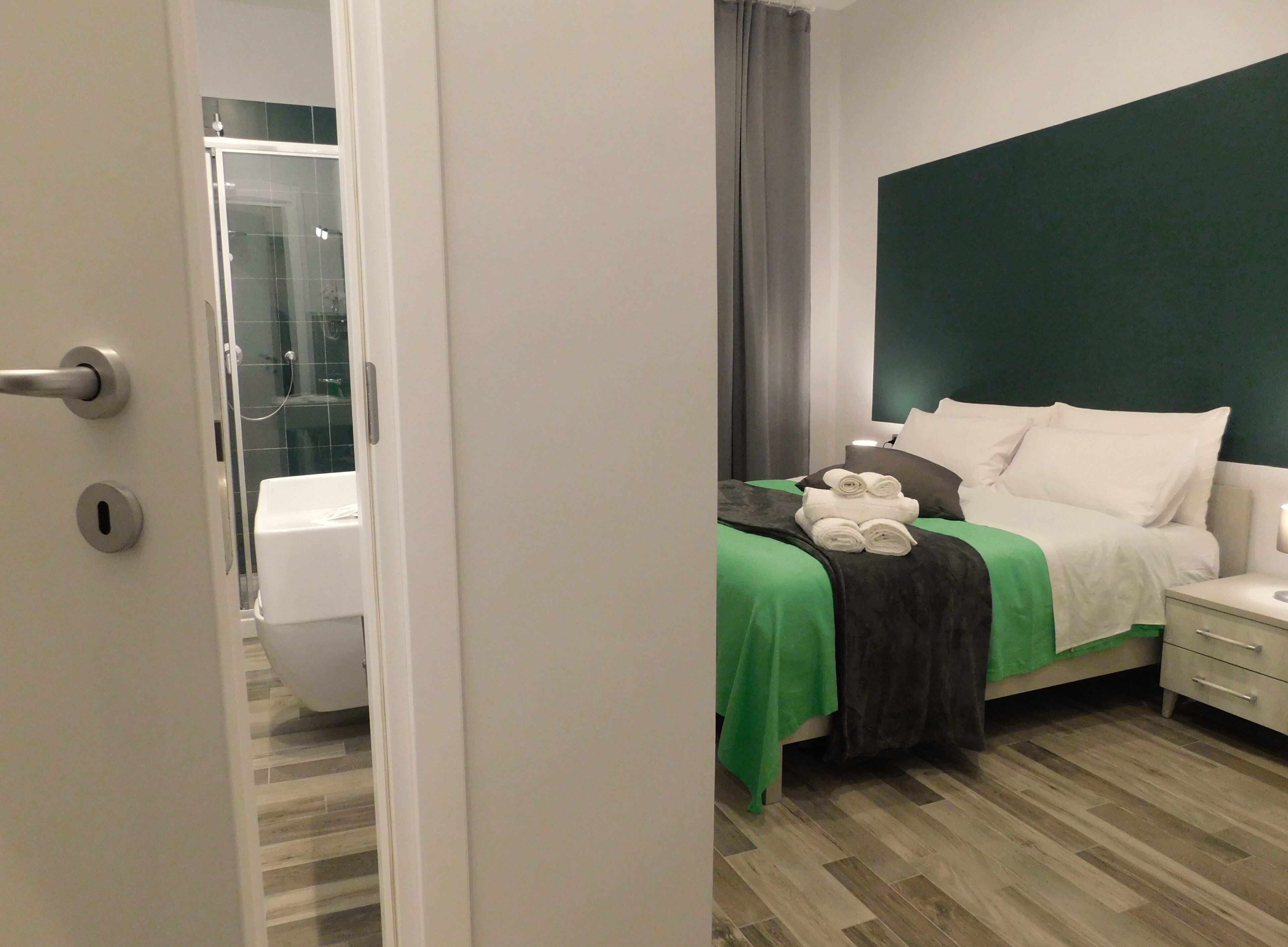 A hotel room with a bed, a bedside table, a nightstand, and a bathroom visible through an open door.