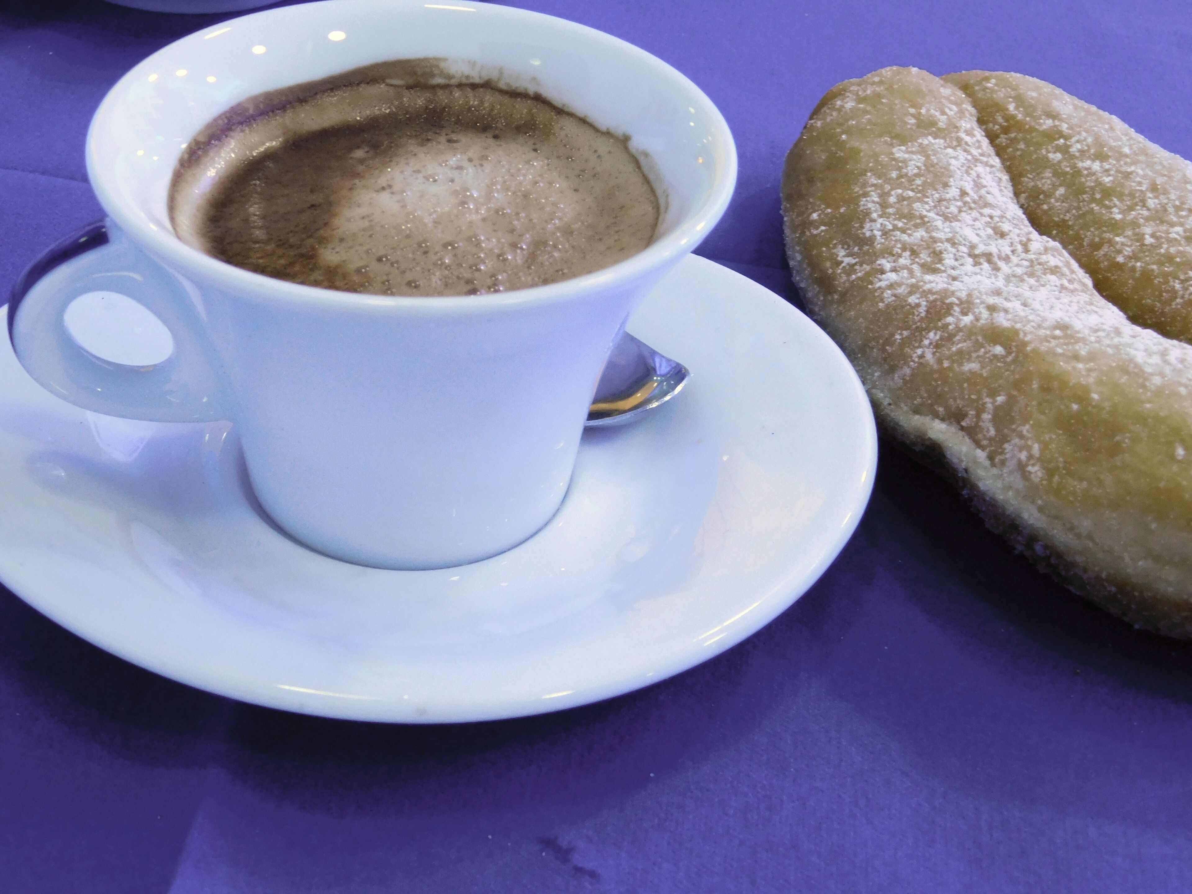 A white cup of coffee with a saucer, a powdered pastry, and a spoon on a purple table.