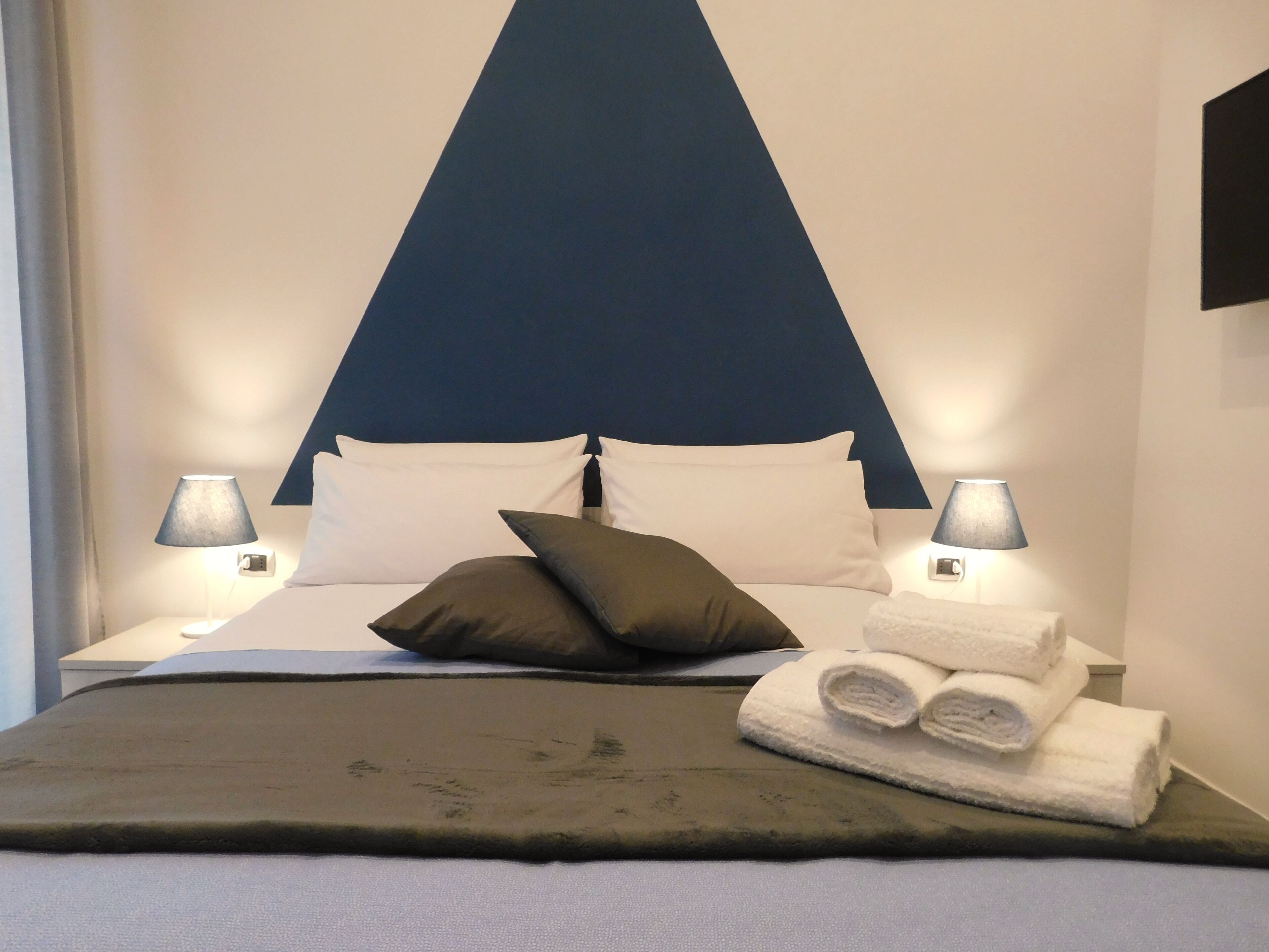 A neatly made bed with a dark green blanket, white pillows, and a dark brown pillow. There are two bedside lamps and a stack of white towels on the side of the bed.