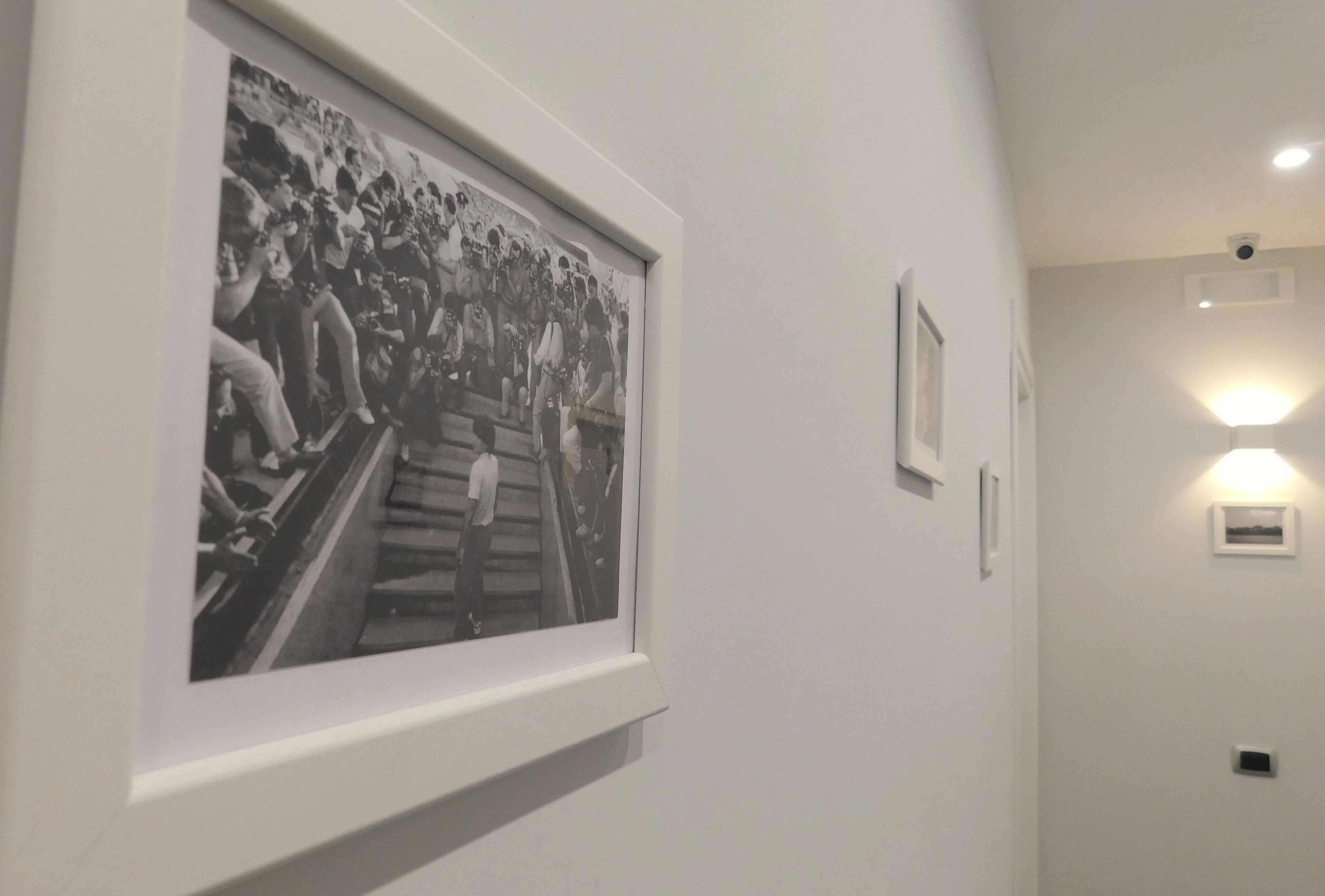 A framed black and white photograph of a crowd on stairs, a wall-mounted light, and a security camera.