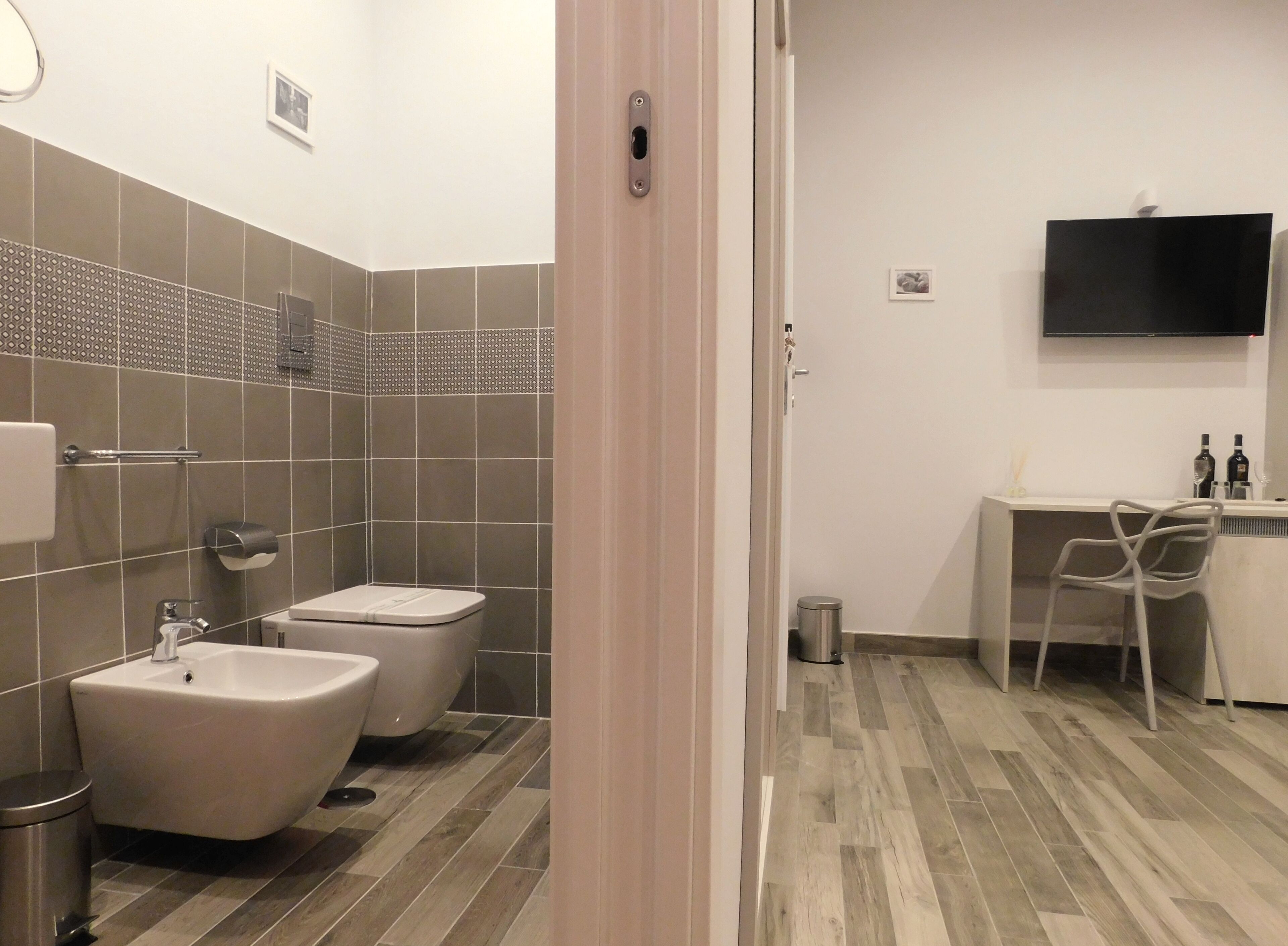 Une salle de bain moderne équipée d’un bidet, de toilettes et d’un lavabo. On y trouve une petite poubelle et une chaise près d’un bureau sur lequel est fixé un téléviseur à écran plat.