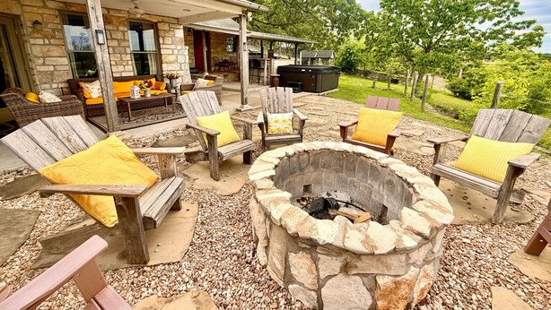 A stone fireplace surrounded by wooden chairs with cushions, a covered patio area with outdoor furniture, and a stone building with a metal roof. - Private Hilltop Farmhouse Incredible Views, Petting Zoo Animals & Hot Tub (Fredericksburg)