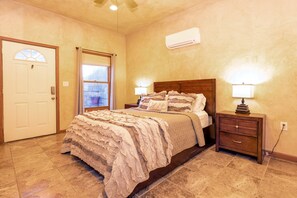 A bedroom with a bed, bedside tables, a nightstand, a window with curtains, an air conditioning unit, and a door.