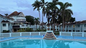 A swimming pool with a tiered water feature, surrounded by palm trees and multi-story buildings with balconies. - The Point, Negril -1 BR, Ocean & beaches, Pool, Tennis, Snorkel 5 star location! (Negril)