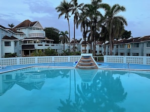 A swimming pool with a tiered water feature, surrounded by palm trees and multi-story buildings with balconies. - The Point, Negril -1 BR, Ocean & beaches, Pool, Tennis, Snorkel 5 star location! (Negril)