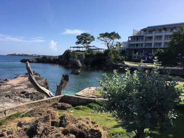 A coastal view with a rocky shoreline, a wooden structure on a cliff, and a building with balconies overlooking the sea. - The Point, Negril -1 BR, Ocean & beaches, Pool, Tennis, Snorkel 5 star location! (Negril)