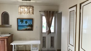 Una habitación con una mesa blanca, sillas, un cuadro de una playa, una lámpara colgante y una puerta con ventanal.