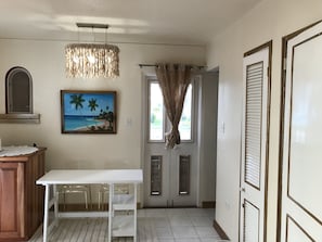 A room with a white table, chairs, a painting of a beach, a hanging light fixture, and a door with a window. - The Point, Negril -1 BR, Ocean & beaches, Pool, Tennis, Snorkel 5 star location! (Negril)