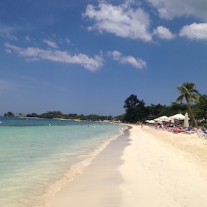 A beach with clear turquoise water, white sand, and palm trees. There are umbrellas and people on the beach. - The Point, Negril -1 BR, Ocean & beaches, Pool, Tennis, Snorkel 5 star location! (Negril)
